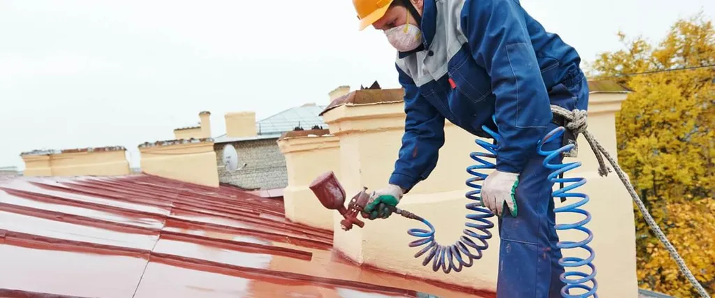 A roofer wearing a hard hat, gloves, and blue coveralls sprays red paint onto a metal roof with a spray gun, connected to a blue air hose, while secured with a safety rope. The background shows chimneys and autumn trees.