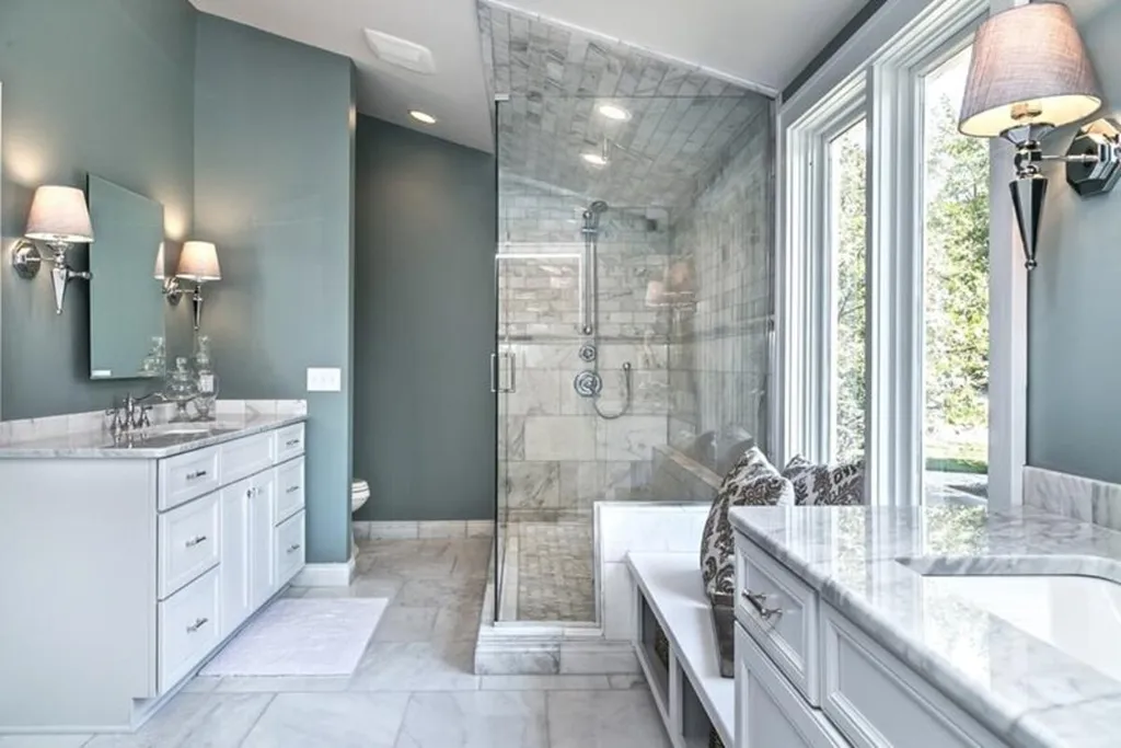 A modern bathroom with a large glass-enclosed marble shower, white double vanity with marble countertop, two wall sconces, a window seat with a patterned cushion, and large windows letting in natural light. The walls are painted a soft teal, and the floor is tiled with white marble.
