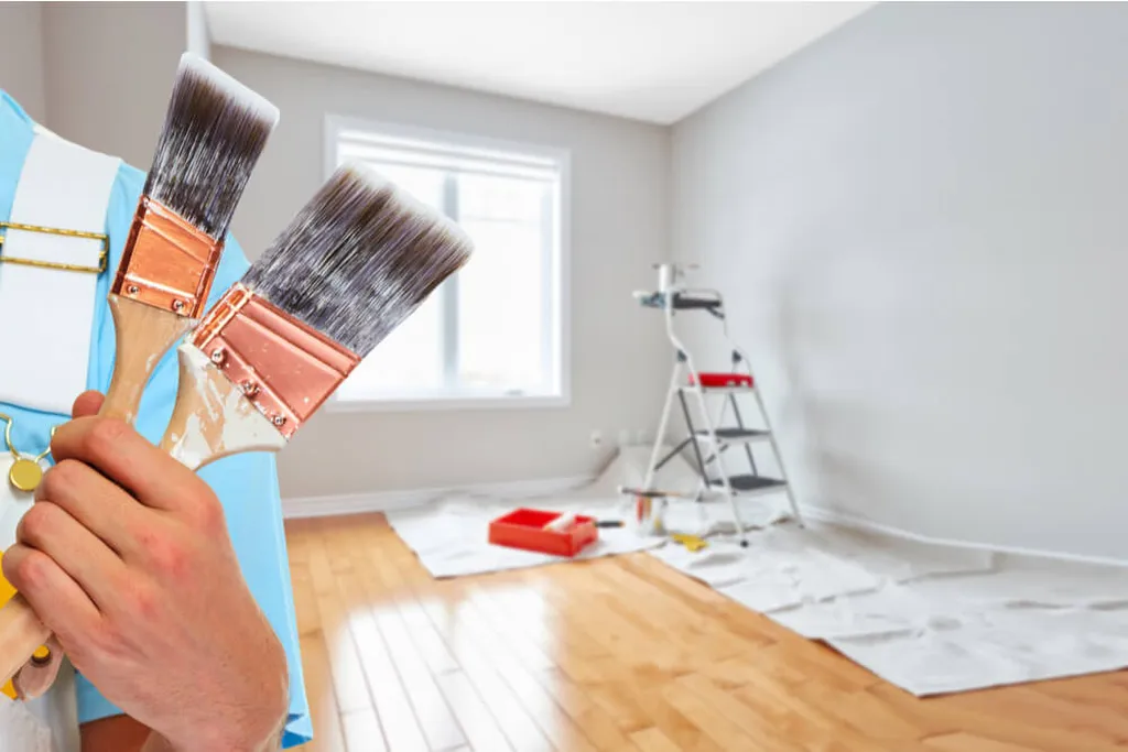 A hand holding two paintbrushes in a room with a ladder, drop cloths, and painting supplies.