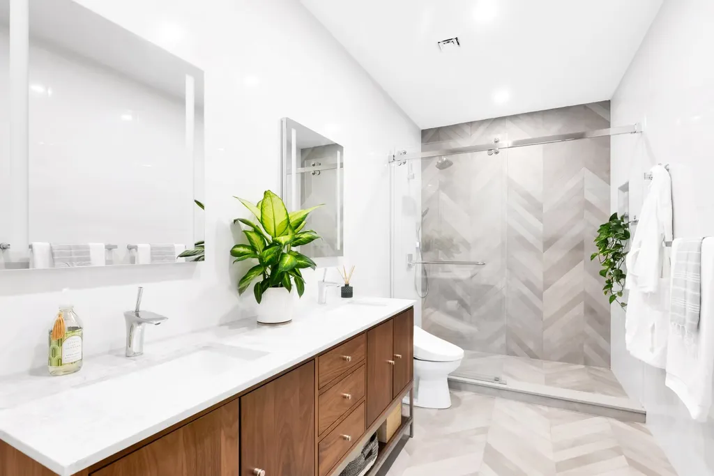 A minimalist bathroom with a wooden vanity, white countertop, modern toilet, glass shower with herringbone tiles, and green plants on the counter and near the shower.