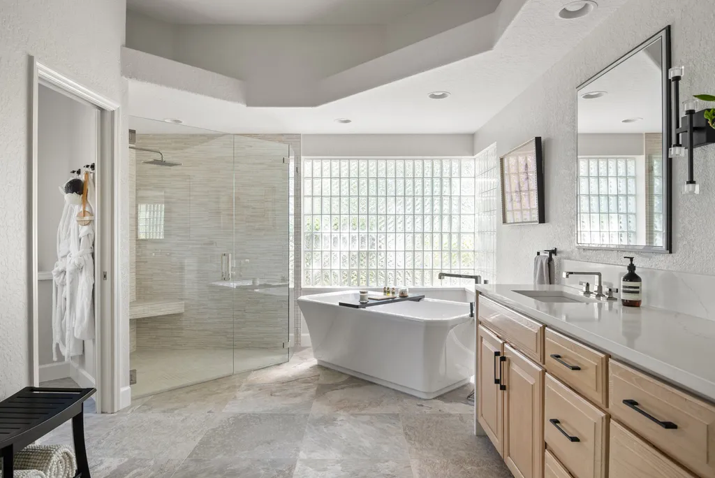 A spacious bathroom featuring a white freestanding bathtub with a black tray, a glass shower with beige tiles, a light wood vanity with a white countertop, and a large mirror. Glass block windows allow natural light to flood the space, while robes hang near the door and a small black bench holds folded towels. The floor is tiled with large gray stone tiles.
