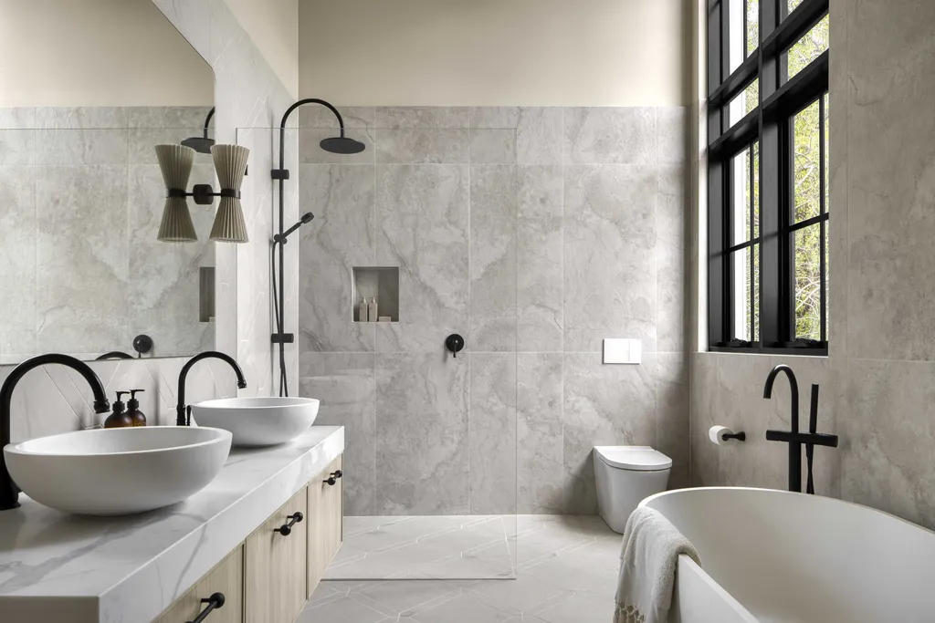 A modern bathroom with a freestanding white tub, double sinks on a light wooden vanity, black faucets, a glass shower, marble walls, and large black-framed windows.