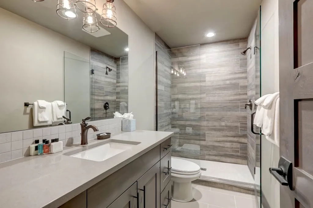 A minimalist bathroom featuring a glass-enclosed shower with gray tiled walls, a gray vanity with a white countertop and sink, a white toilet, and a tissue box on the counter. Two white towels hang on a rack, and a towel is draped over the shower door.