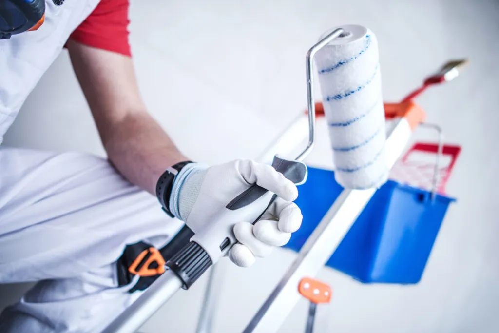 A close-up of a painter's hands in white gloves holding a paint roller, with a blue tray and other painting tools on a ladder in the background. The painter wears white overalls and a red shirt.