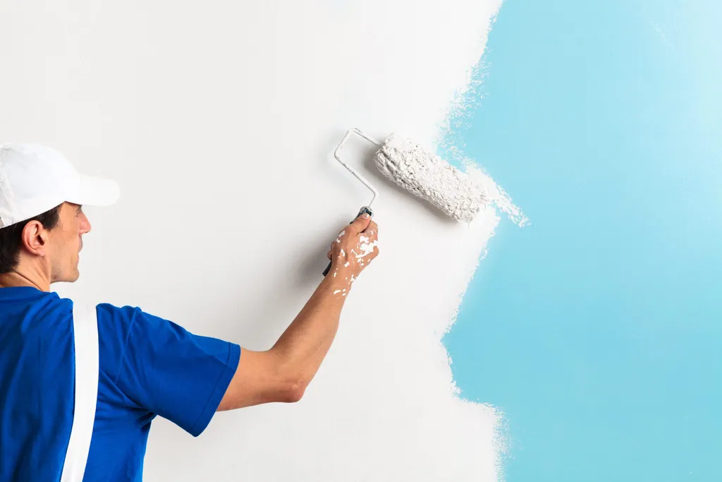 A person in a white cap and blue shirt painting a wall with a roller, applying light blue paint.