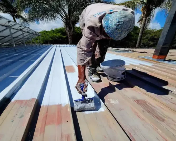A roofer in a protective suit and headscarf paints a roof white with a roller, using a can of paint nearby. The scene includes palm trees, solar panels, and a clear blue sky in the background.