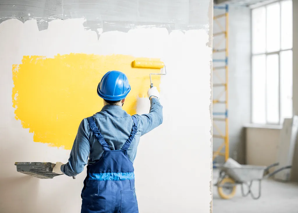 A person in a blue hard hat, denim shirt, and blue overalls stands in a construction site, painting a wall yellow with a roller. A tray of paint is in one hand, with scaffolding, a wheelbarrow, and large windows in the background.