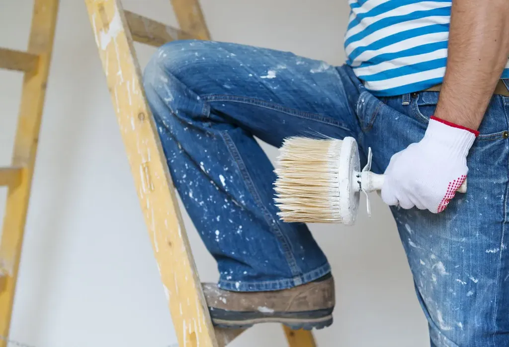 A person in a striped shirt and jeans, holding a paintbrush on a ladder, with paint splattered on their clothes.