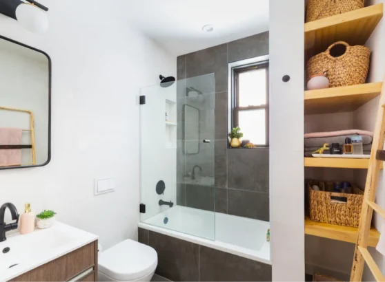 Small modern bathroom with bathtub, glass screen, wooden shelving, and black fixtures