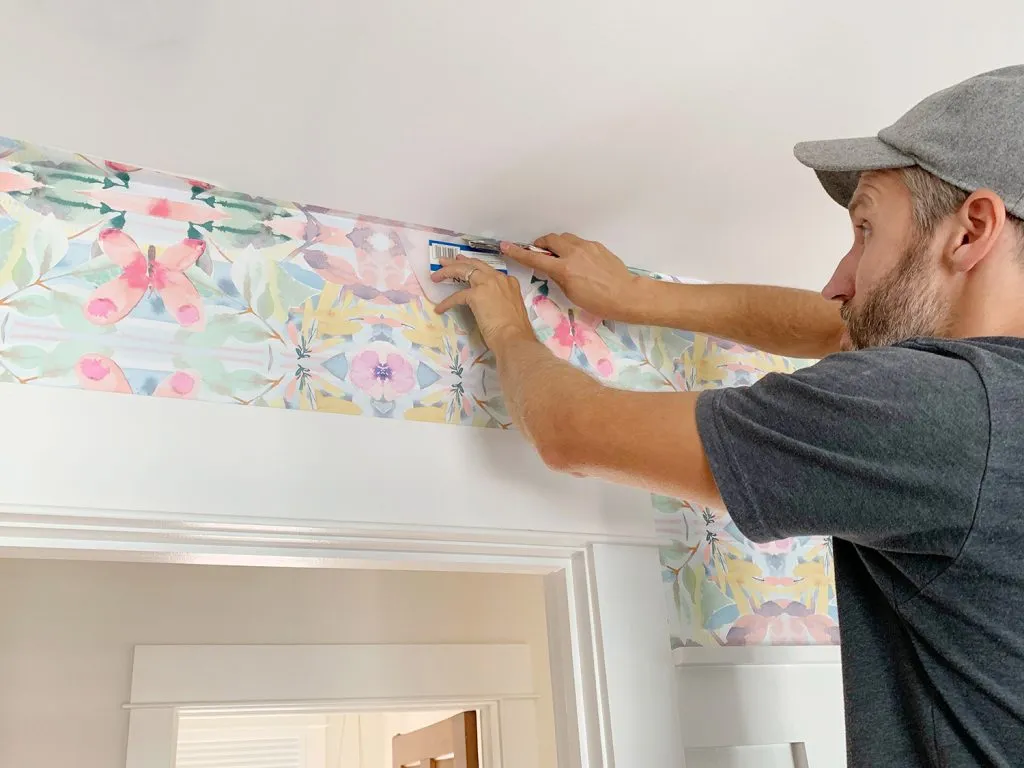 A person in a gray cap and dark shirt carefully installs a floral wallpaper border with a butterfly and flower pattern along the top edge of a wall, using a utility knife to trim the excess.