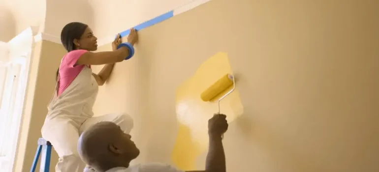 Two painters in a room, one taping the wall with blue painter's tape on a ladder, while the other rolls yellow paint onto the wall, demonstrating careful interior painting.