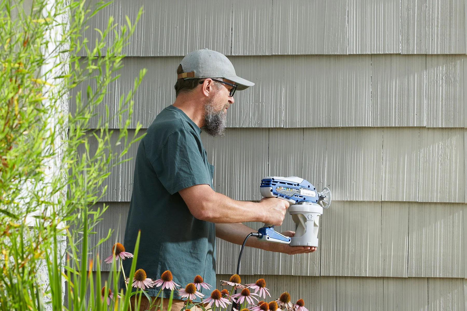 A man in a green shirt and cap holding a power washer near a gray house wall, with flowers in the foreground.