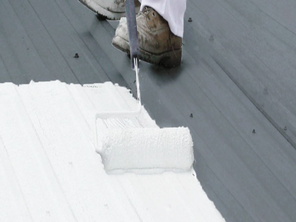 A person in white overalls and boots using a roller to paint a wooden deck white.