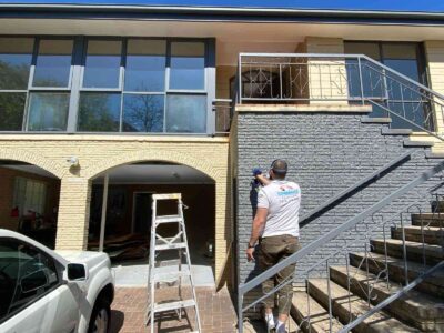 Painter applying grey paint to textured exterior wall in Fadden