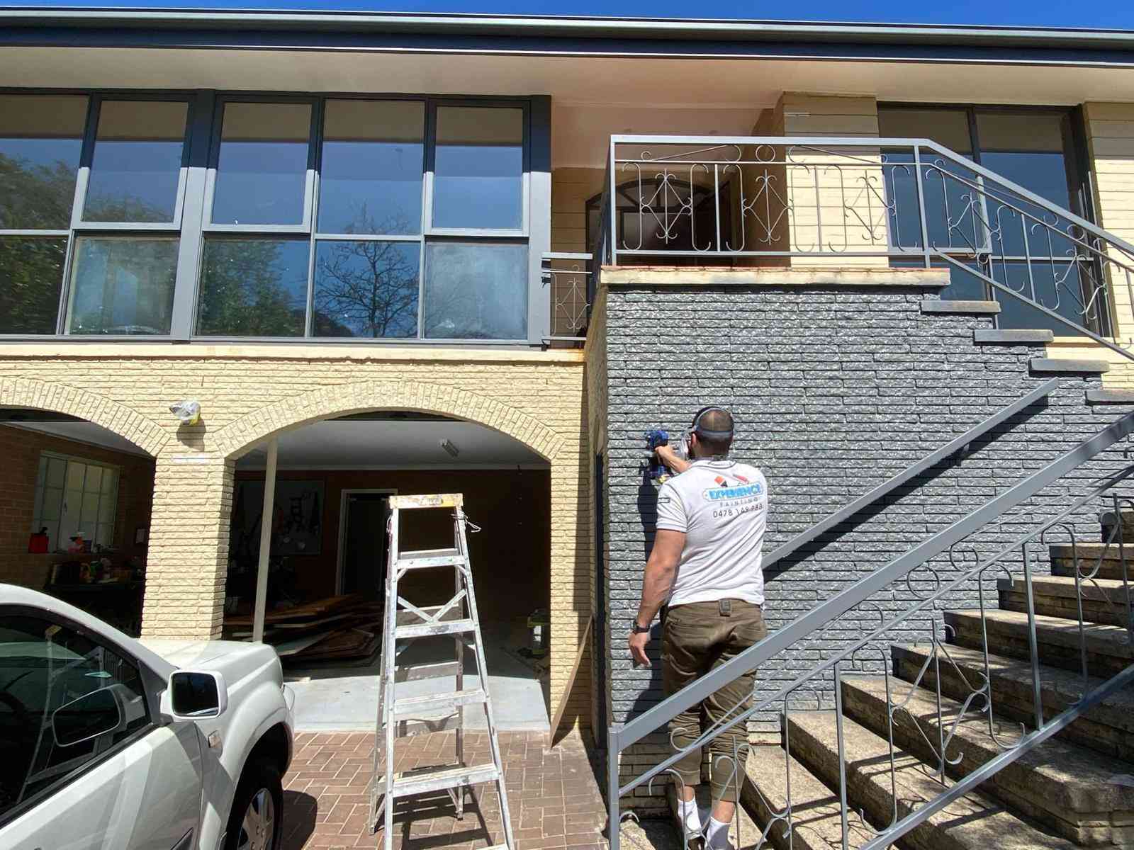 Painter applying grey paint to textured exterior wall in Fadden
